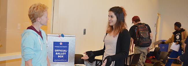 Two female student election workers speak to each other and smile next to a ballot box inside a voting location.